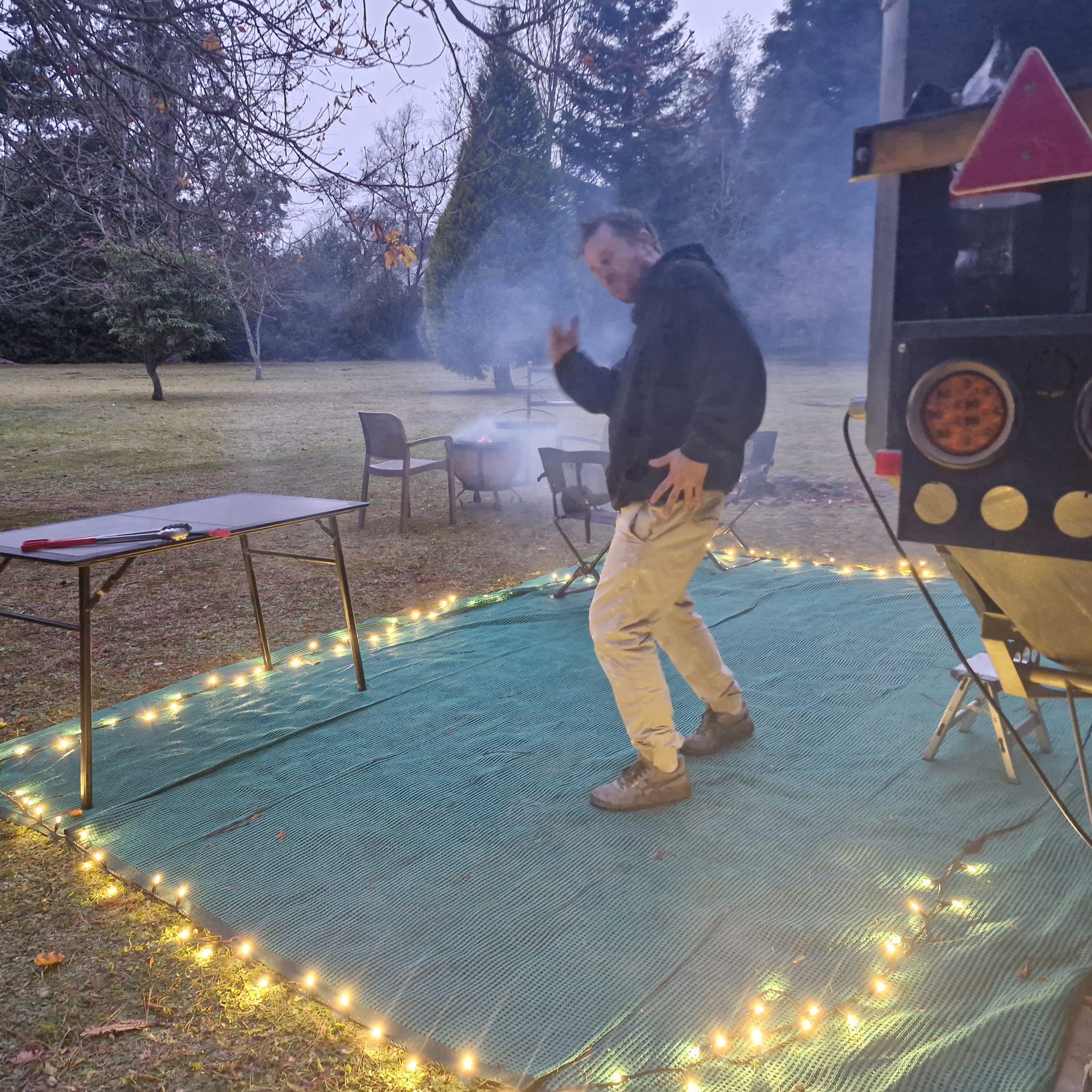 A camper crossing the groundsheet at dusk beside an off-road trailer at Maylodge campsite, frost on the lawn