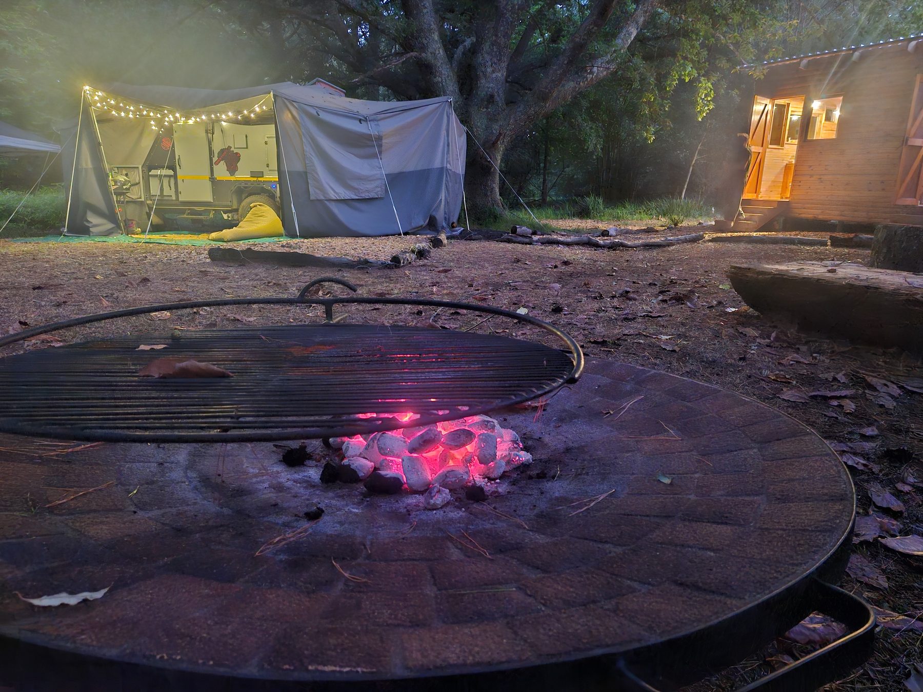 Campfire coals glowing at dusk at Maylodge campsite in Hogsback, with a lit tent and cottage behind