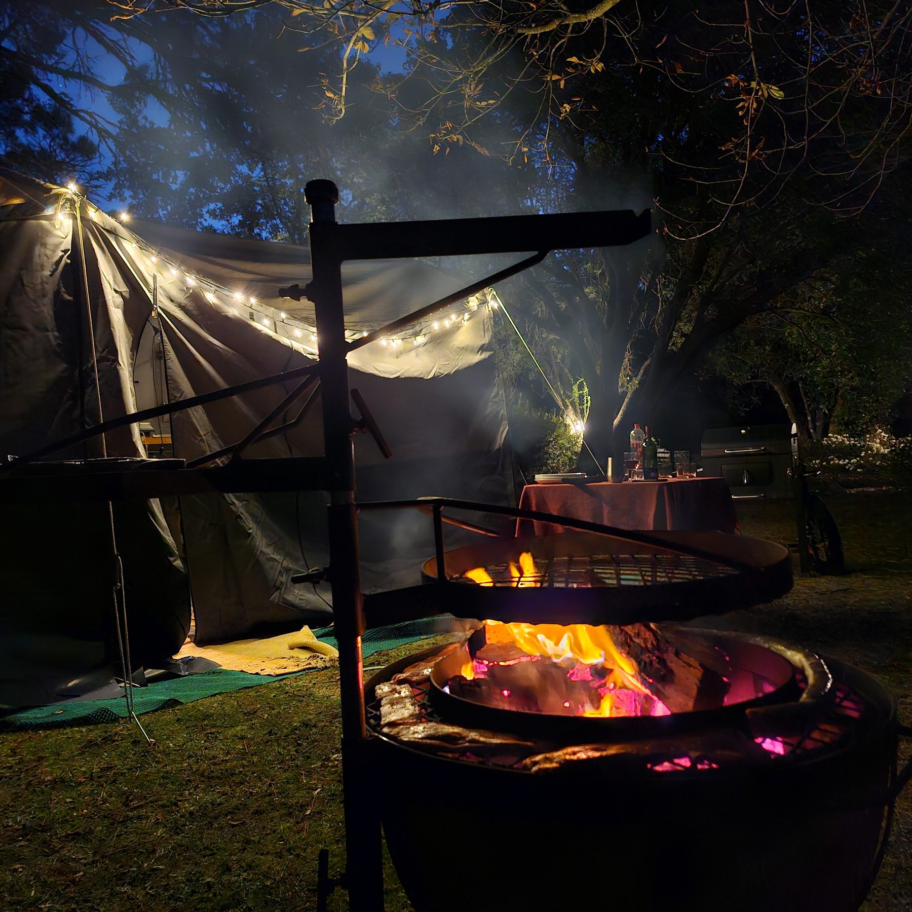 A fire pit burning at night in front of a canvas trailer tent strung with fairy lights at Maylodge campsite
